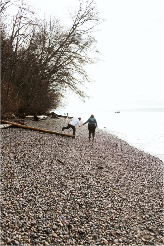 An engaged couple holding hands and walking along the beach during their Seahurst Park engagement session