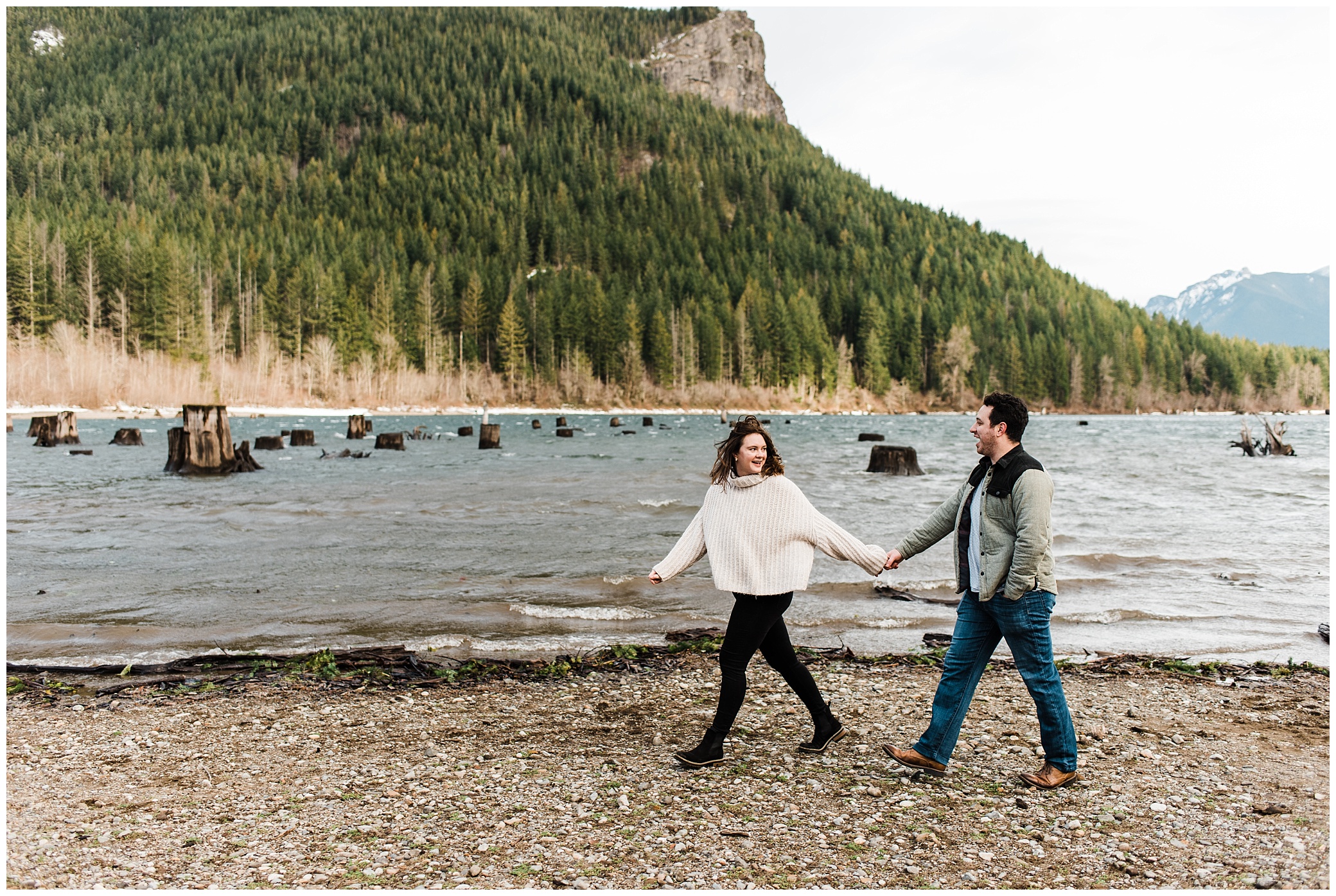An engaged couple walks along a lake, holding hands and looking at each other