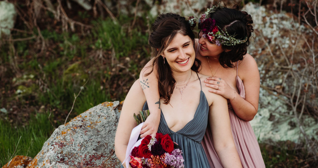 Two marriers smiling and cuddling each other, celebrating their queer wedding. One is wearing a blue dress. The other is wearing a pale pink dress and flower crown.