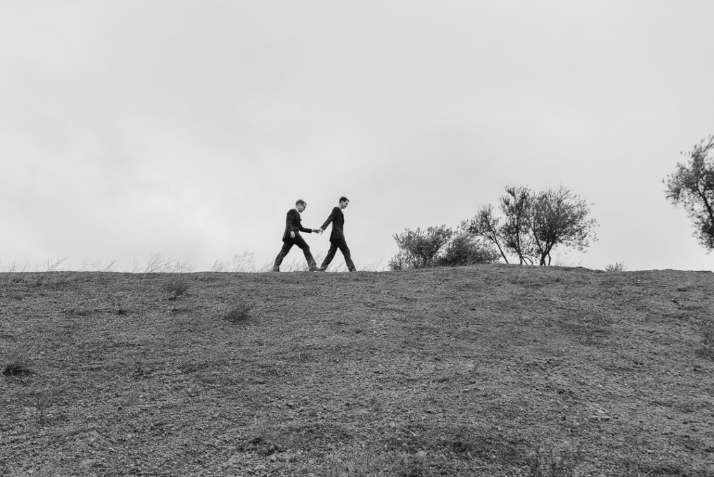 Two marriers in suits walk across a ridge, holding hands.