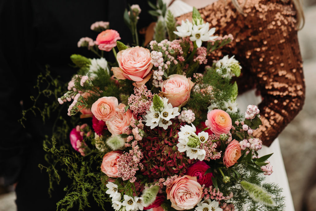 A large bouquet of greenery and pink, white, and maroon flowers is held by a bride in a sparkling copper jacket.