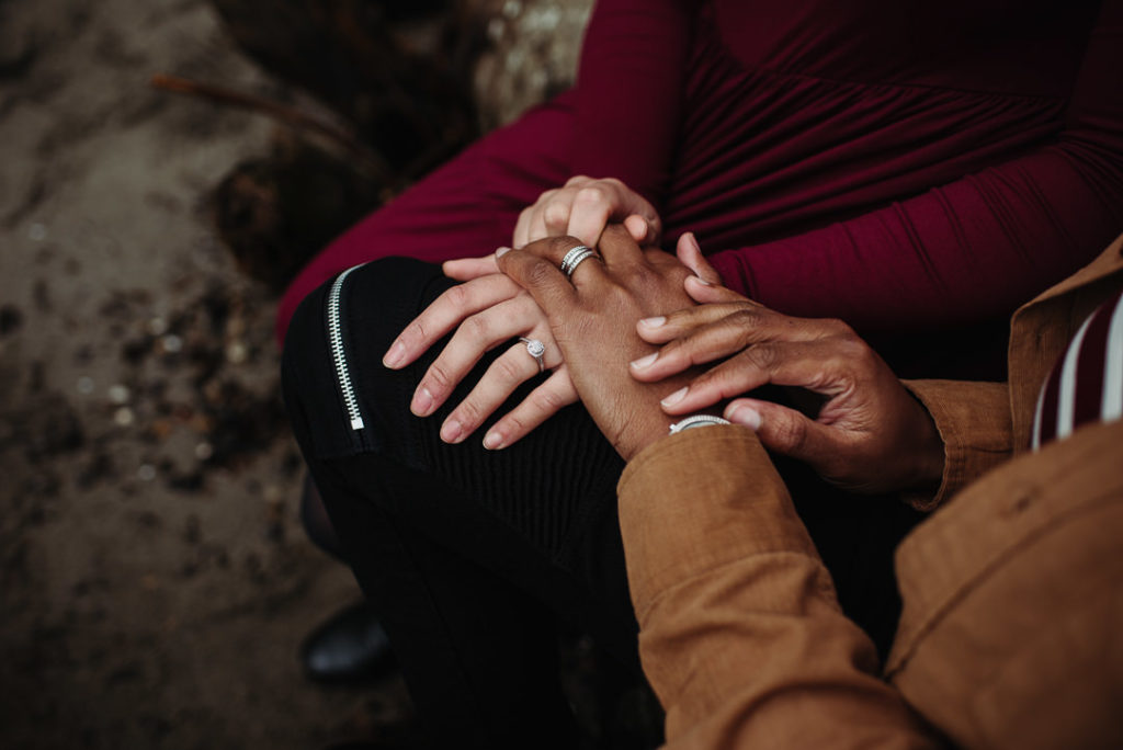 An engaged couple sitting together with their hands overlapping in their laps. Both are wearing engagement rings.