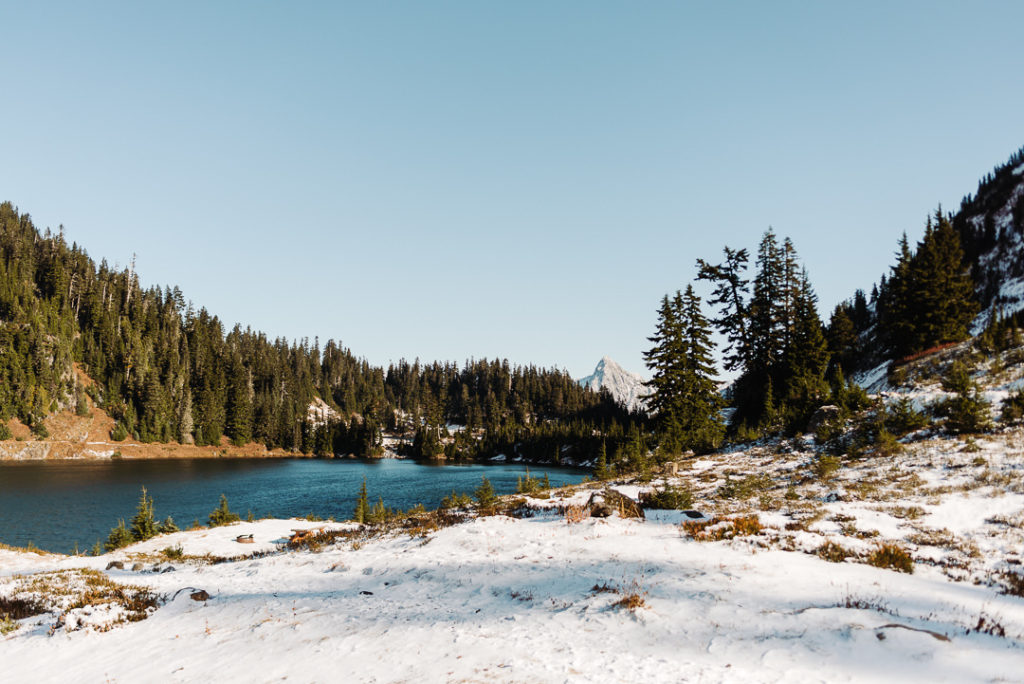 A snowy landscape with evergreen trees, a blue lake, and a mountain rising in the background.