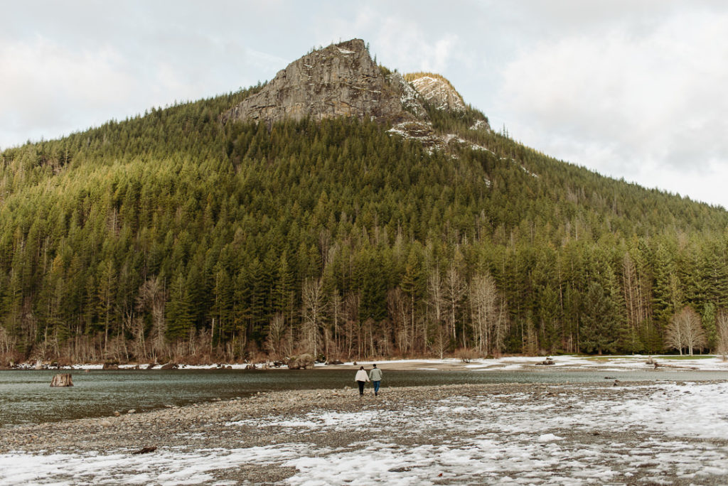 A couple walks away in the distance toward Rattlesnake Lake. A forested mountain rises up in front of them.