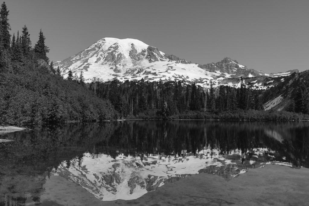 Mount Rainier rising above a forest in the background. A lake in the foreground shows a reflection of the mountain.