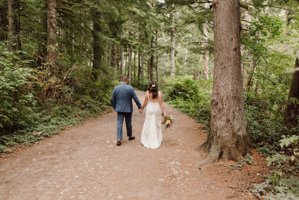 Two marriers walk away from the camera down a forest path, surrounded by trees. They are holding hands, and one is holding a bouquet.